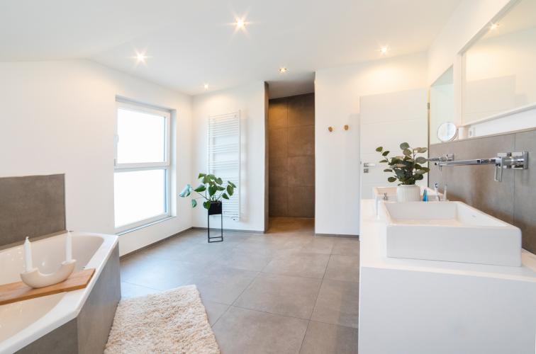 Das Badezimmer zeigt sich elegant und schlicht. Weiß dominiert den Raum und harmoniert wunderbar mit den großformatigen Fliesen aus graubraunem Feinsteinzeug.<i> Foto: HELMA Eigenheimbau AG / Ralph Thiele</i>